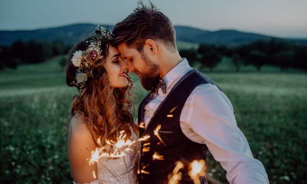 A bride and groom standing in a field at sunset with sparklers